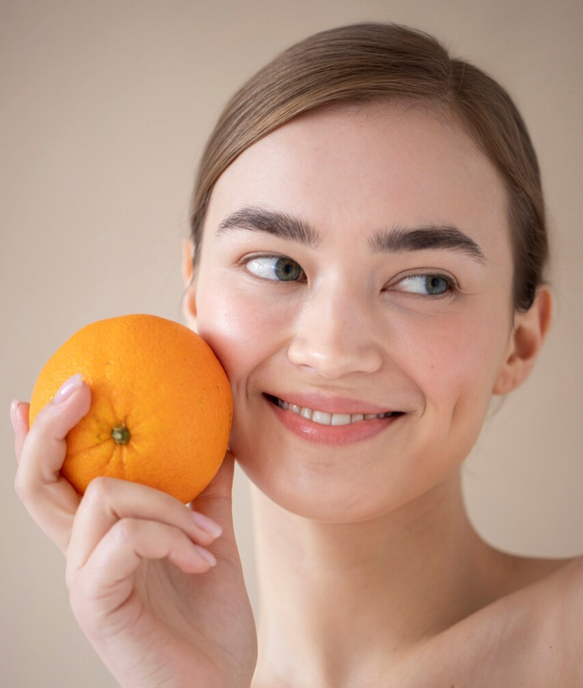 mujer joven con pelo recogido primer plano mirando hacia el lado sonriendo sujeta una naranja junto a su mejilla