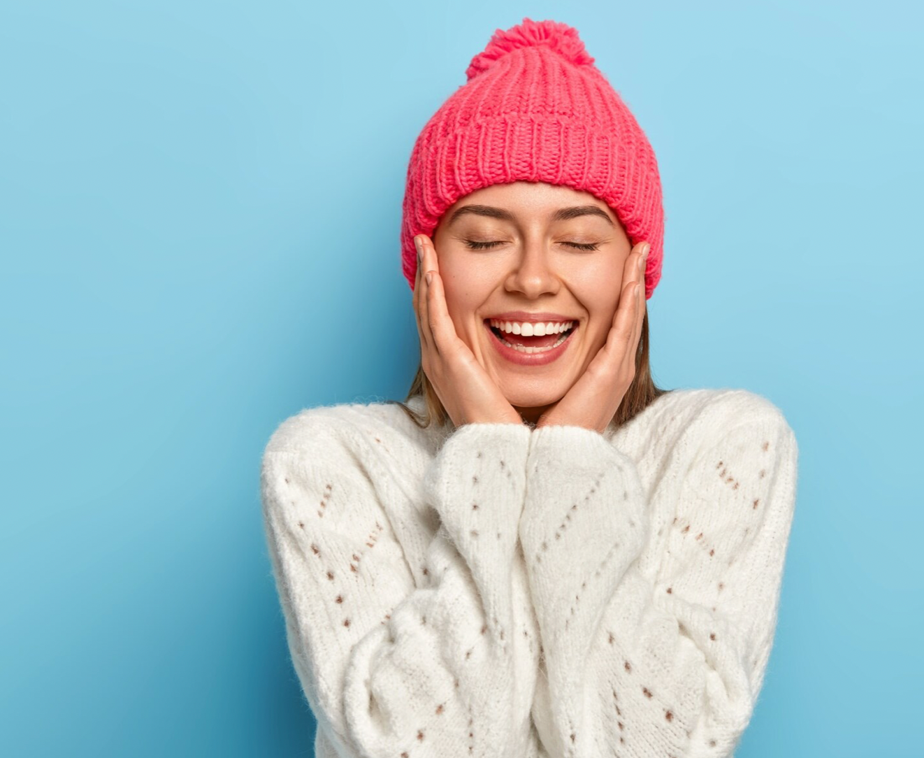 mujer joven con ropa de invierno gorro de lana rosa con pompón y jersery de lana blanco ojos cerrados sonríe con una mano en cada mejilla fondo claro
